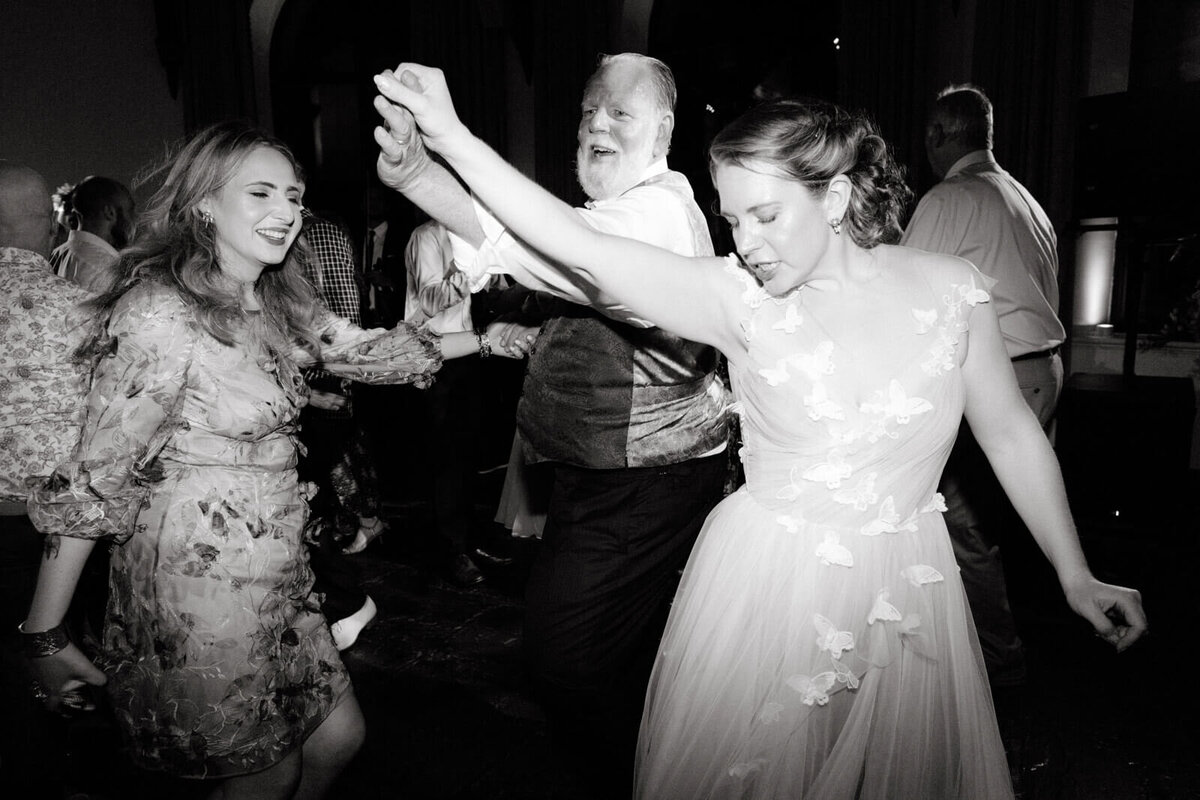 A black and white photo of the bride, dancing with an old man and a woman, with guests in the background