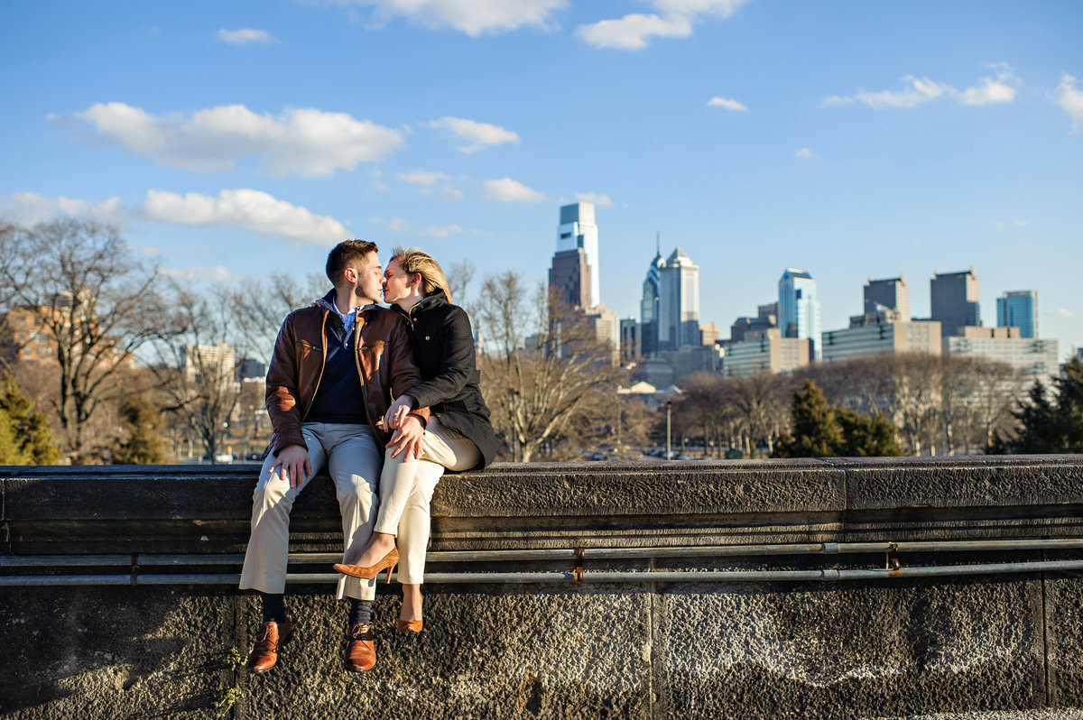 A couple in love sit on the wall of the Philadelphia Art Museum with the center city skyline behind them.