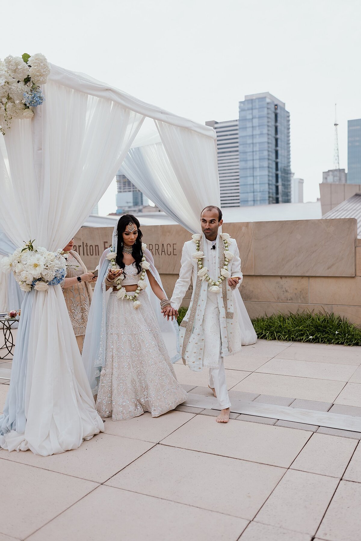 Indian bride and groom walk the seven circles around their white and light blue mandap while wearing white varmala garlands with the Nashville skyline in the background.