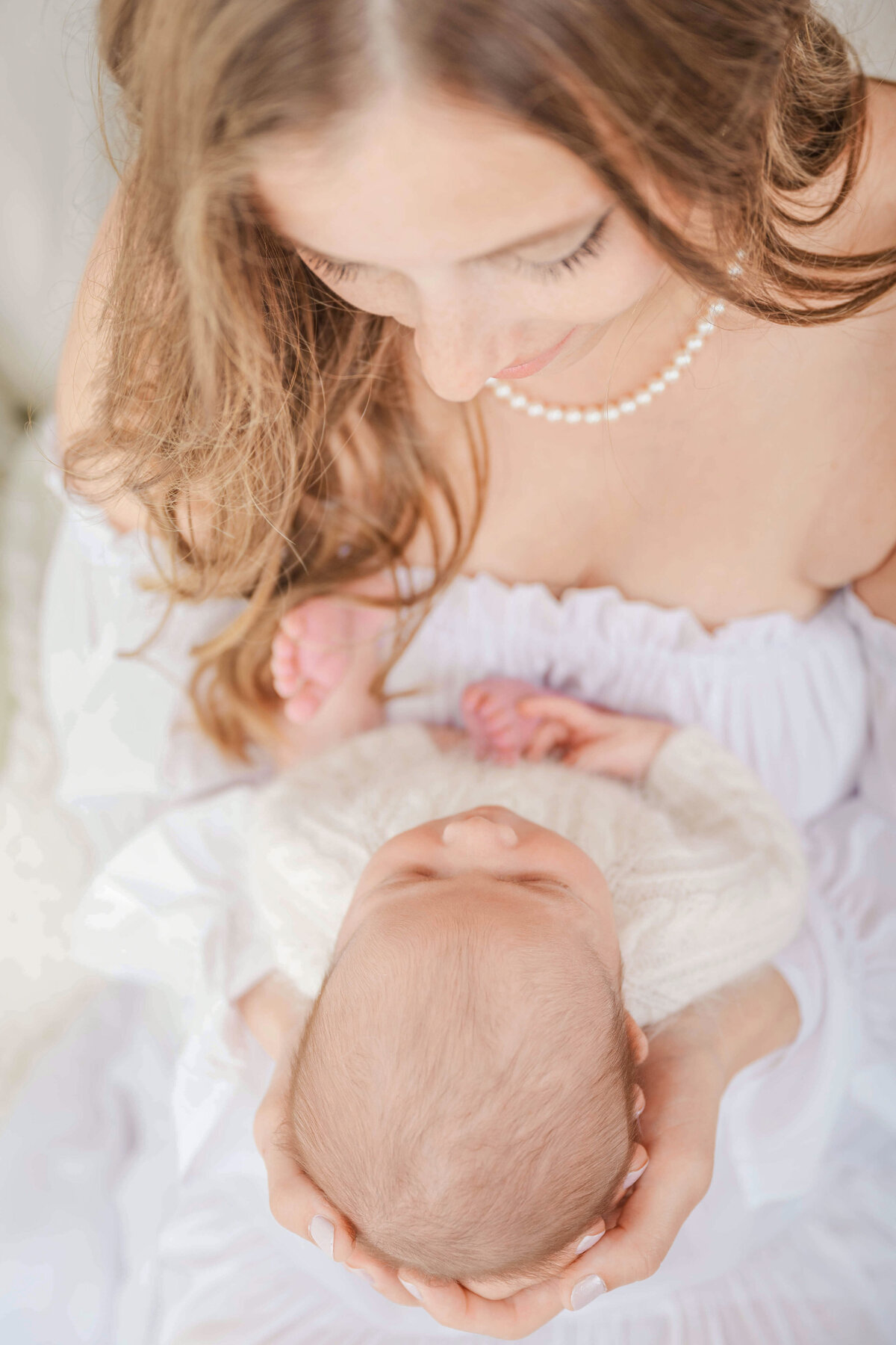 A beautiful woman wearing pearls and looking down at her newborn baby in her lap.