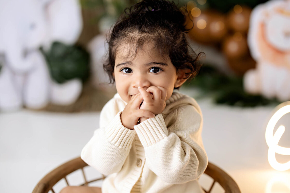 A young child with dark hair and big brown eyes sits on a chair, wearing a cream-colored sweater and smiling shyly with hands near their mouth. Plush animal toys and soft lighting are in the blurred background, evoking a family photographer’s gentle touch.