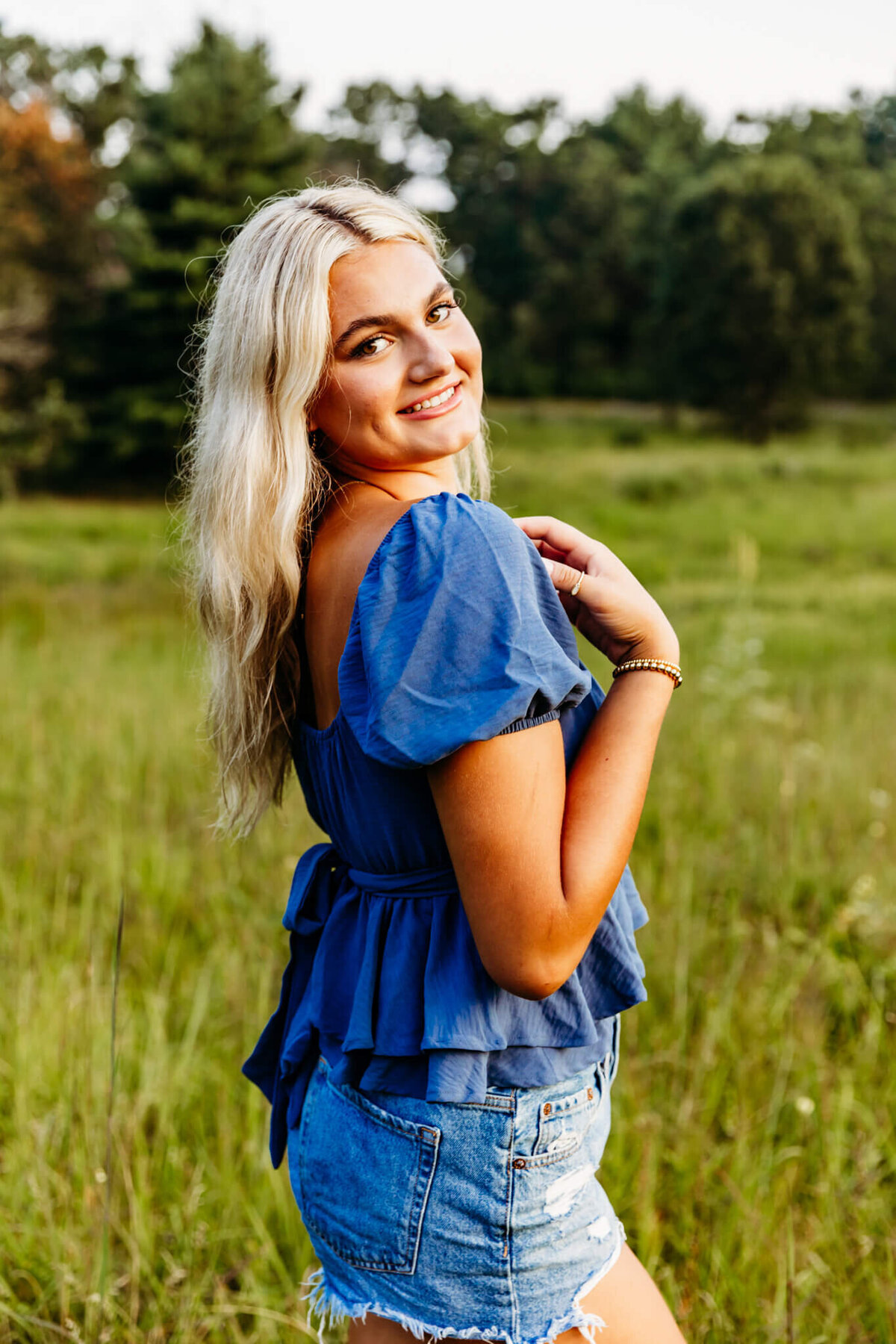 senior girl in a blue short sleeve top standing sideways as she rests her hand on her shoulder