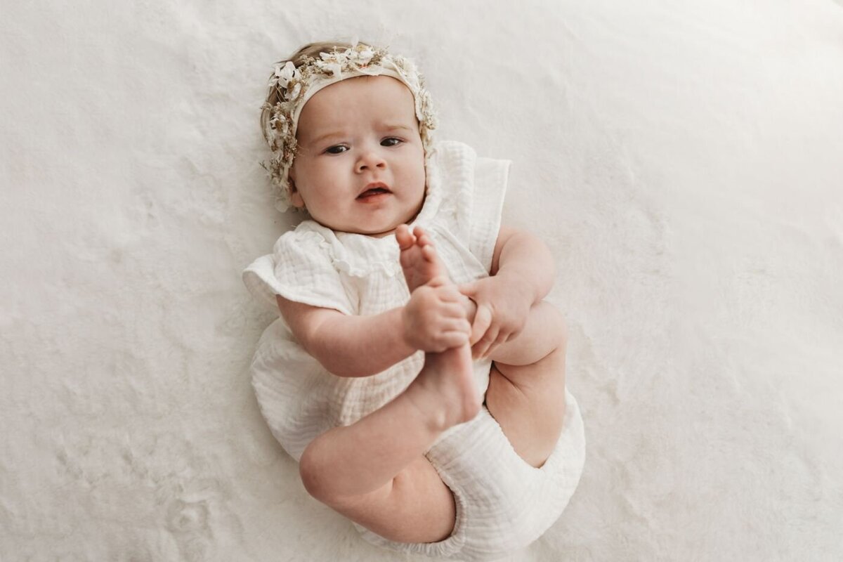 Top-down view of an adorable newborn baby girl wearing a charming floral and greenery crown, playfully holding her feet while lying on a cozy white shag rug