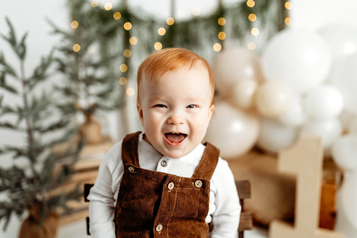 A smiling baby with red hair wearing a brown overalls and white shirt sits in front of a festive background with balloons and greenery. Twinkling lights add a warm ambiance.