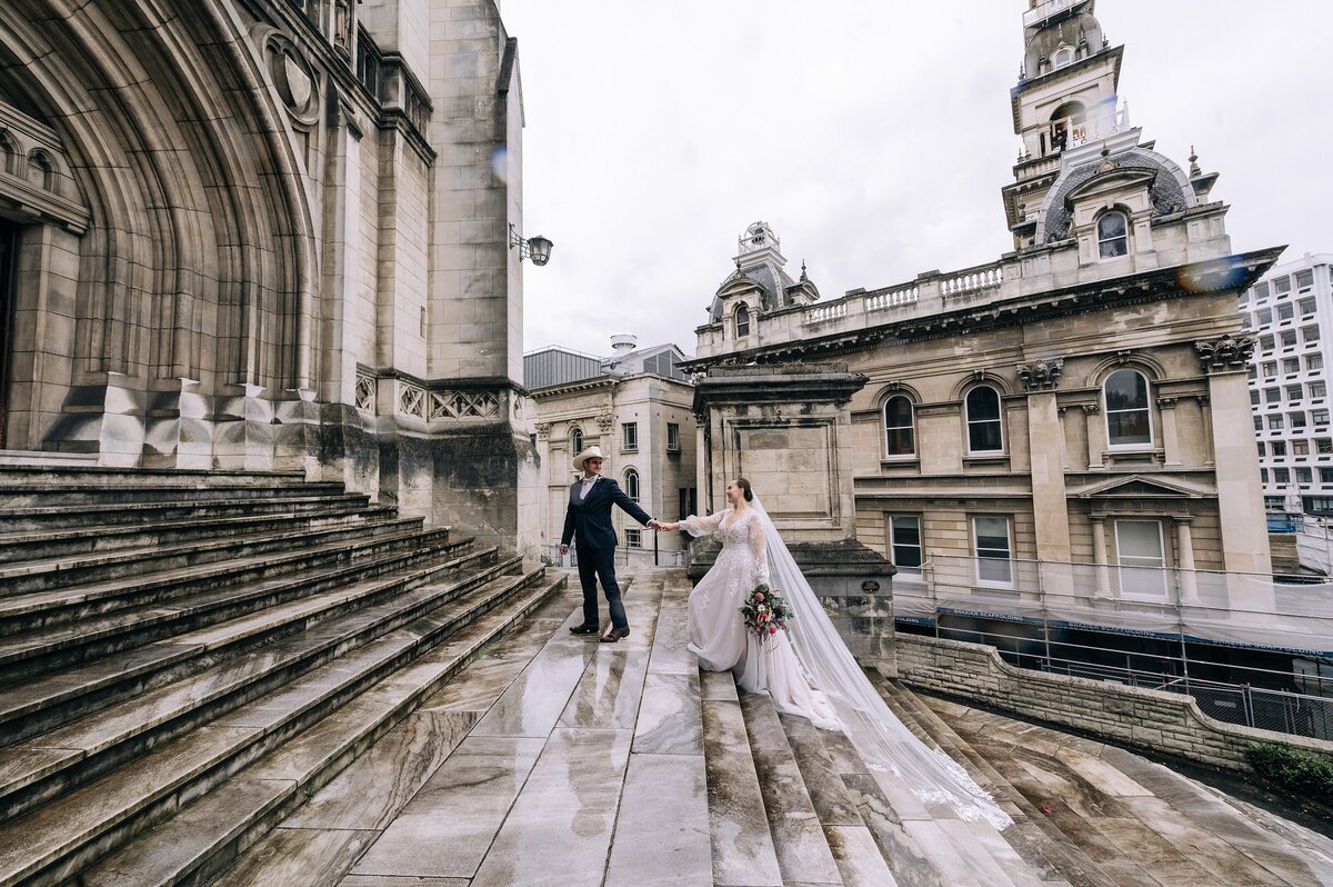 bride and groom walk up the steps of st pauls cathedral in dunedins octagon with a long veil