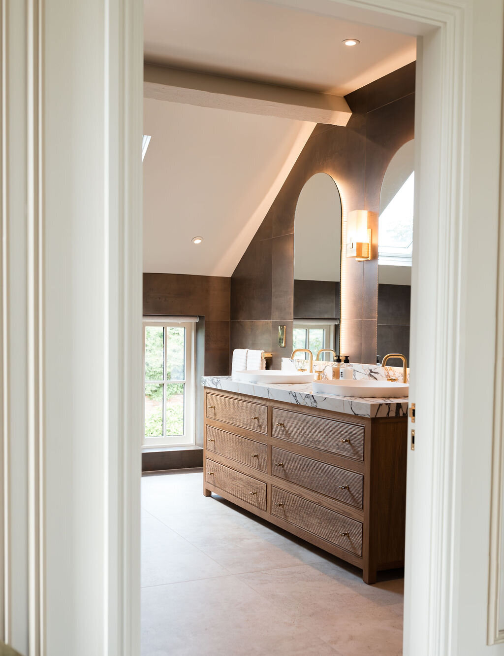 A modern bathroom with a sloped ceiling, featuring a double sink vanity with marble countertops and wooden cabinets. Two arched mirrors are mounted above the sinks, and large windows provide natural light. The room is finished with dark tiled walls and light tiled floors.