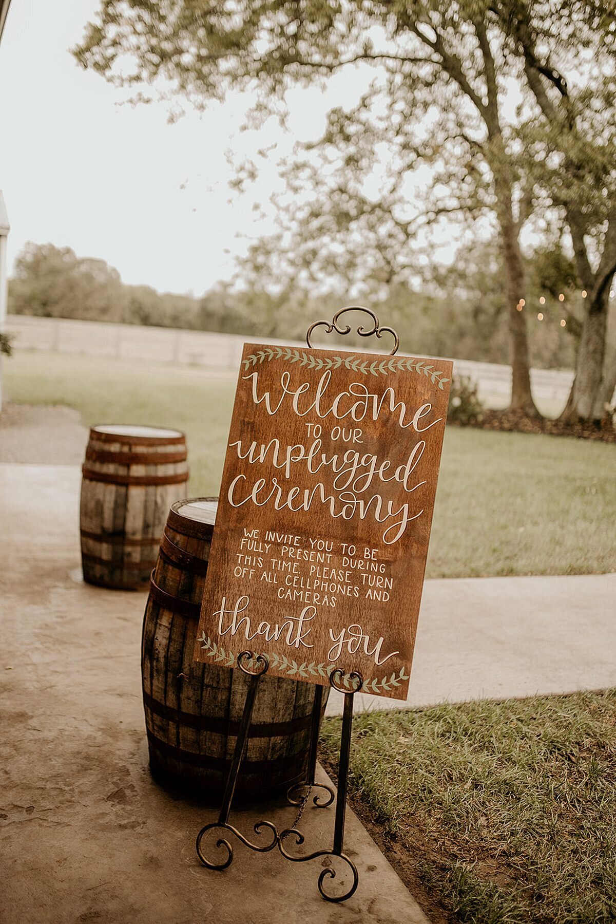 Wood wedding sign made by Lucky Shadow Shop details an unplugged wedding ceremony on a cast iron easel beside two wine barrels.