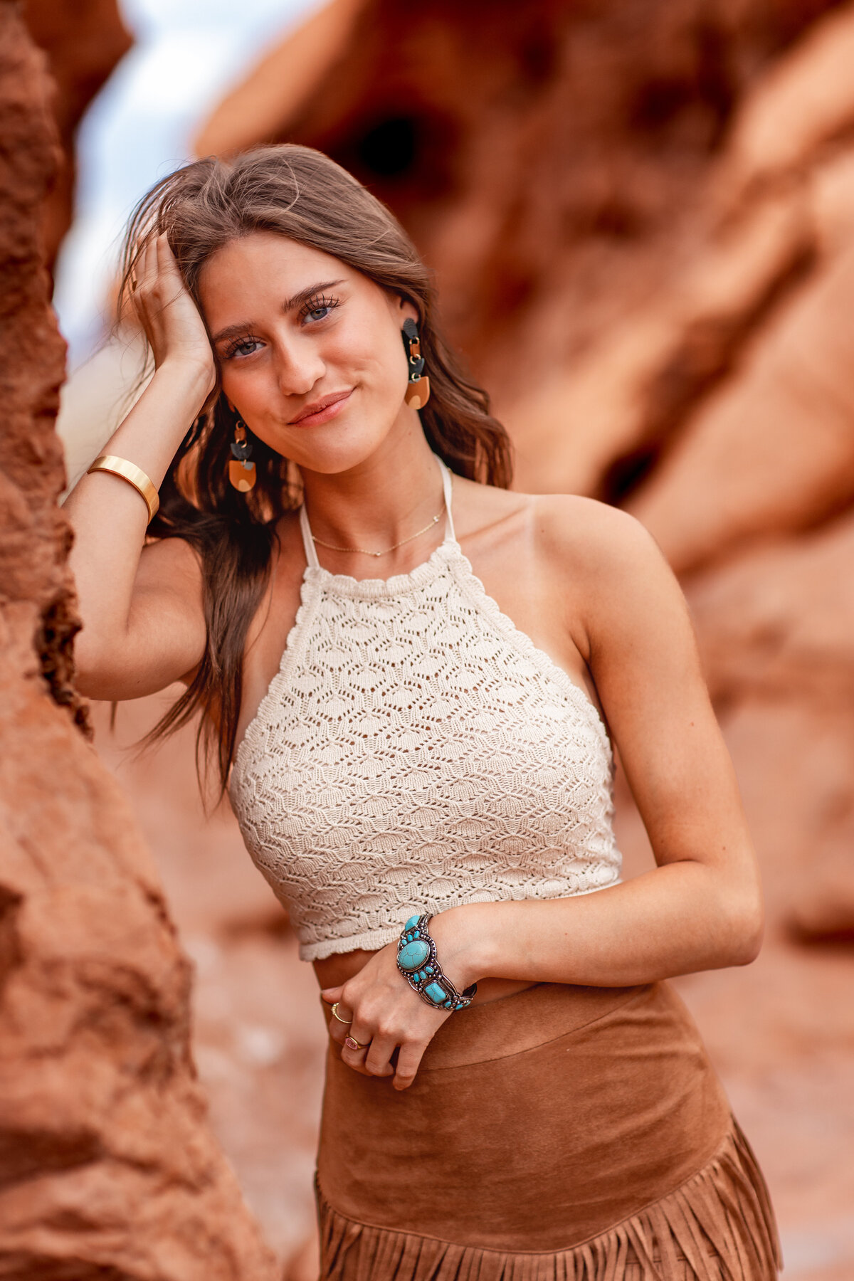 A high school senior with long brown hair leans casually against the red rocks of the Southwest desert, smiling in a textured halter top and turquoise bracelet.  Captured by Brooke Tucker Photography, this boho-inspired senior portrait radiates warmth, natural beauty, and carefree confidence.