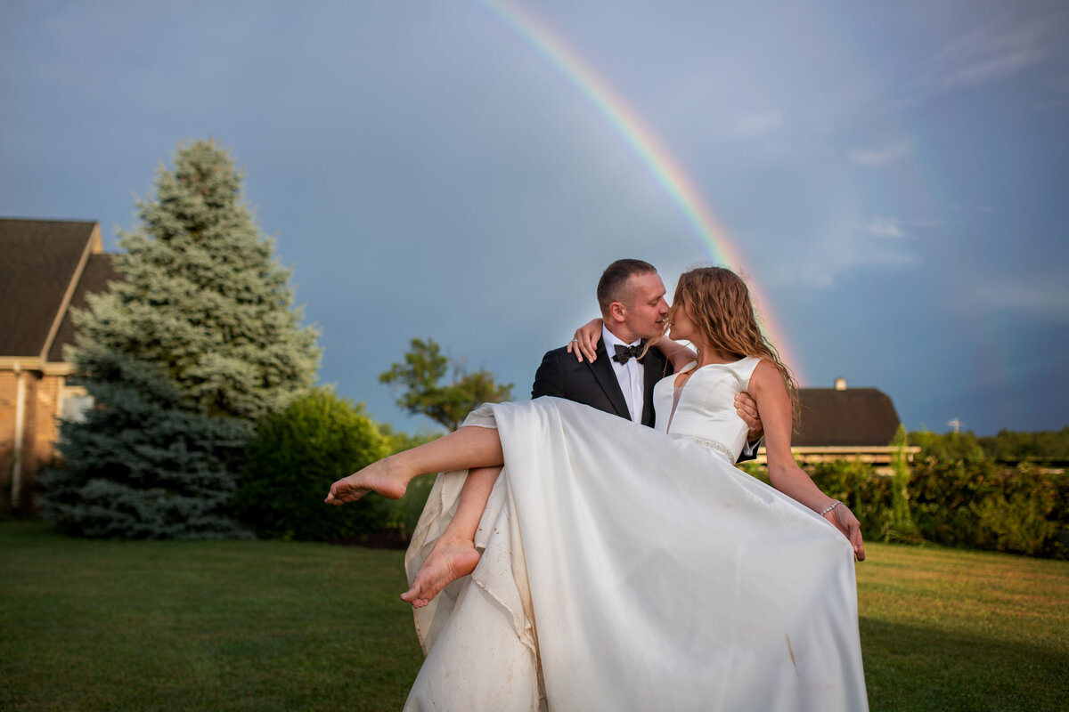 Groom picking the bride up and kissing  her with rainbow in the background behind them