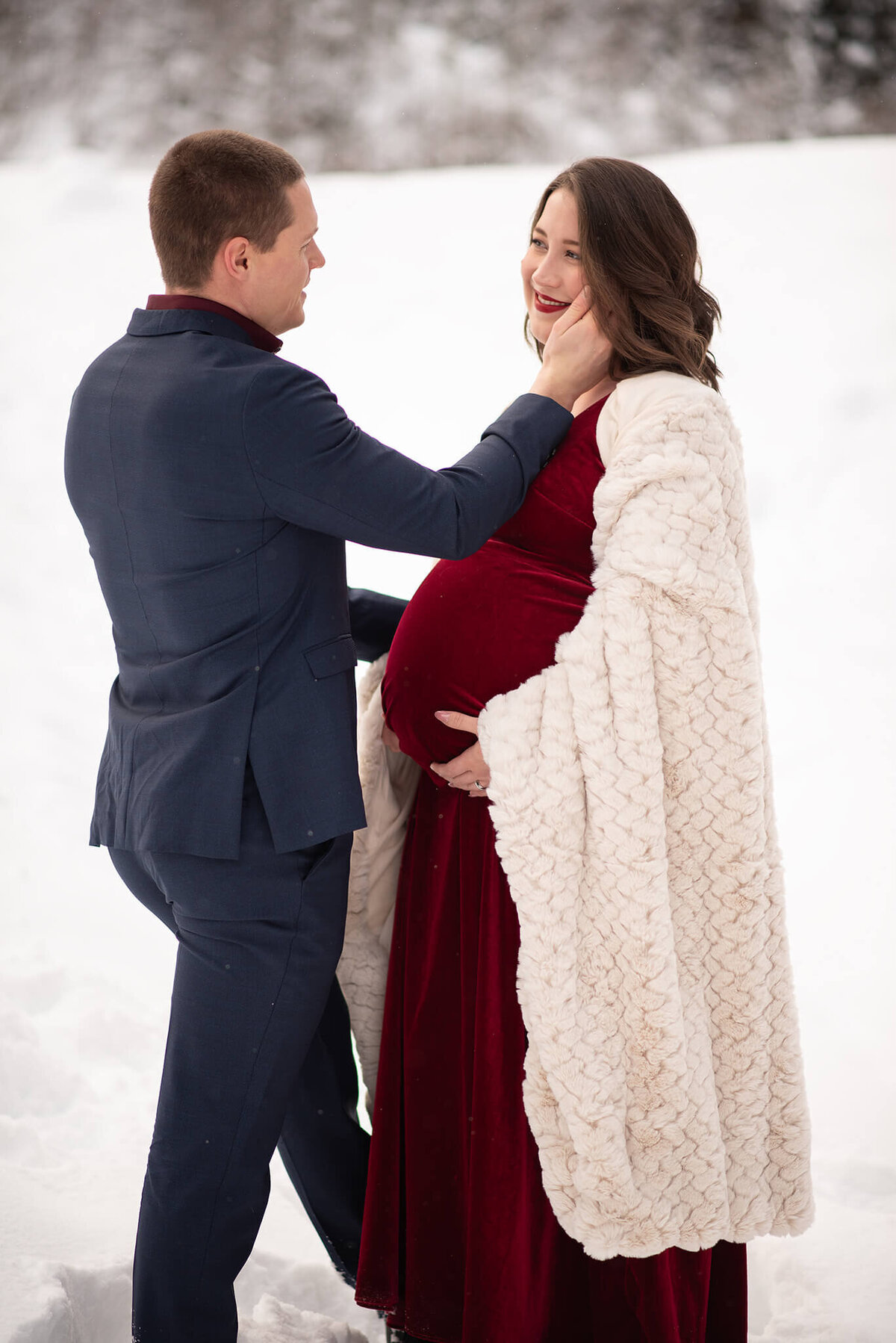 Brittany Danielle captures this image of a husband and wife in Sun Peaks, BC. The pregnant wife wears a red dress under a cozy blanket and the husband in a suit caresses her.