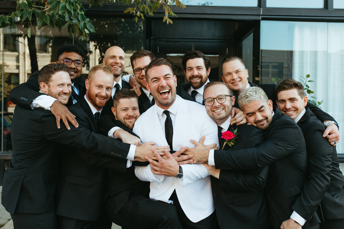 Group of joyful men in formal attire hugging and laughing around the groom in an urban setting