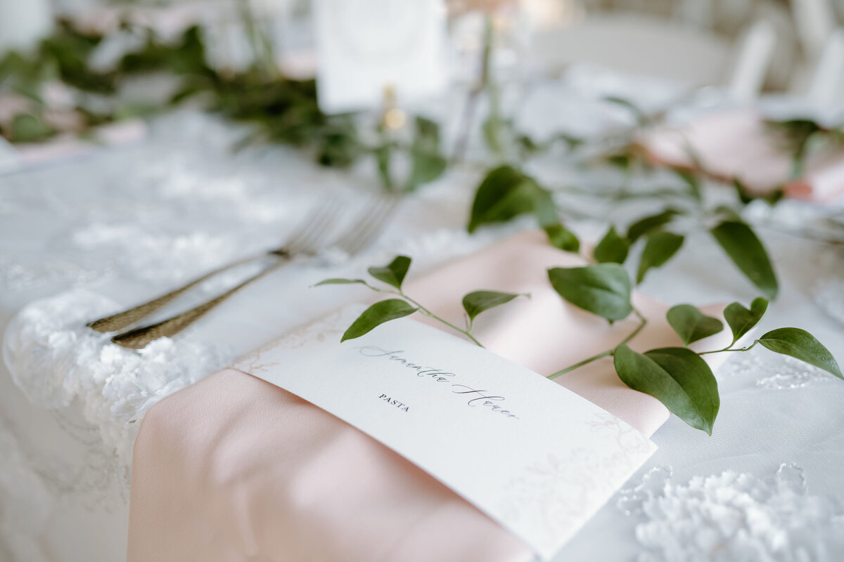 Elegant wedding place card with guest name, styled atop blush pink napkin and greenery; luxury wedding stationery detail.