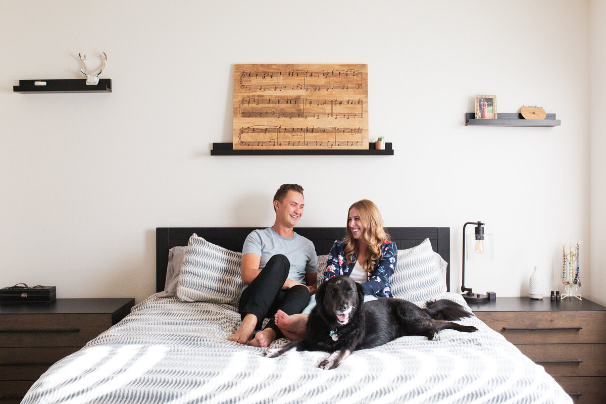 A young husband and wife sit on their bed with their big black lab.
