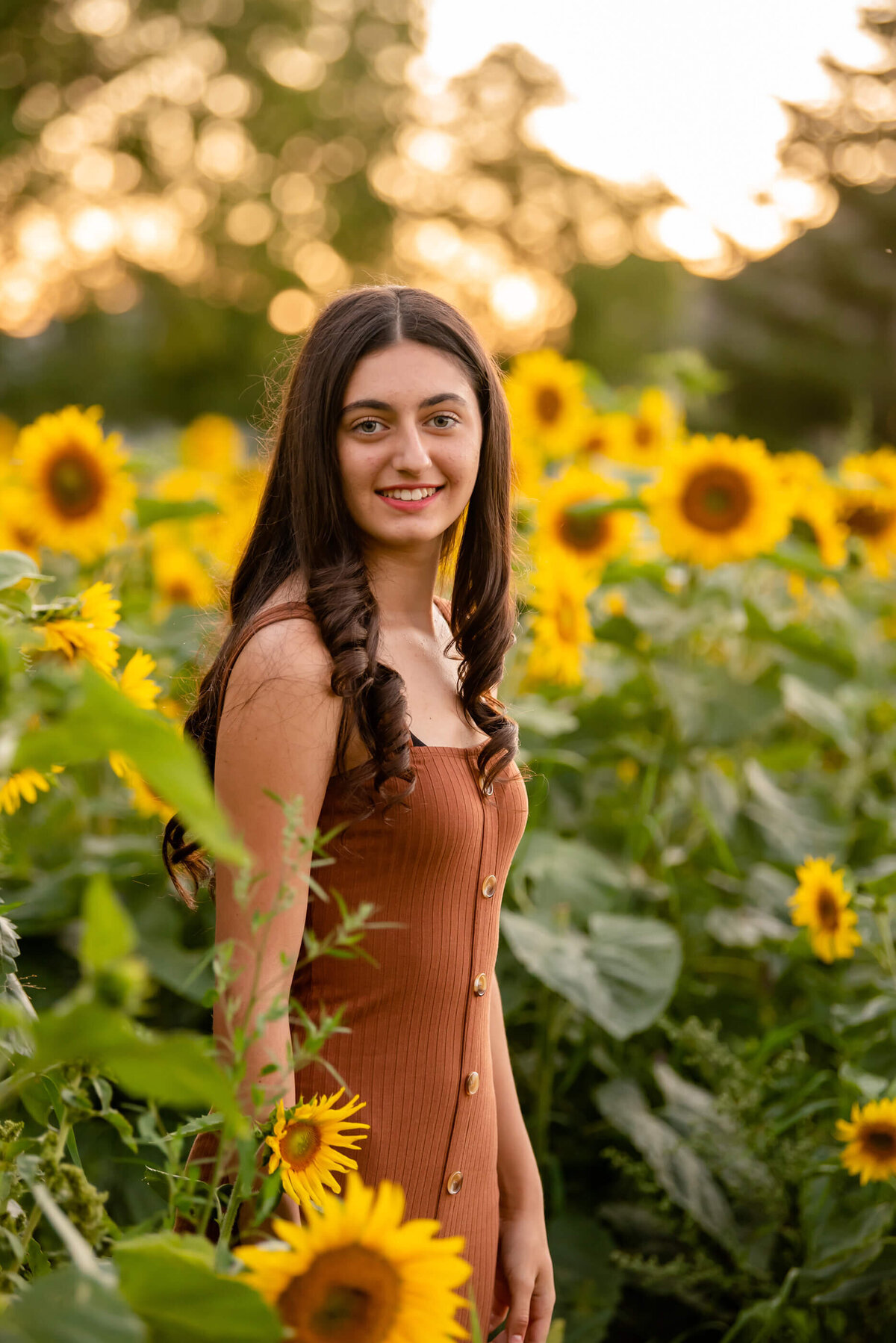 Teen girl with brown hair wearing a burnt orange dress standing in a sunflower field for her family photos in Burlington, On