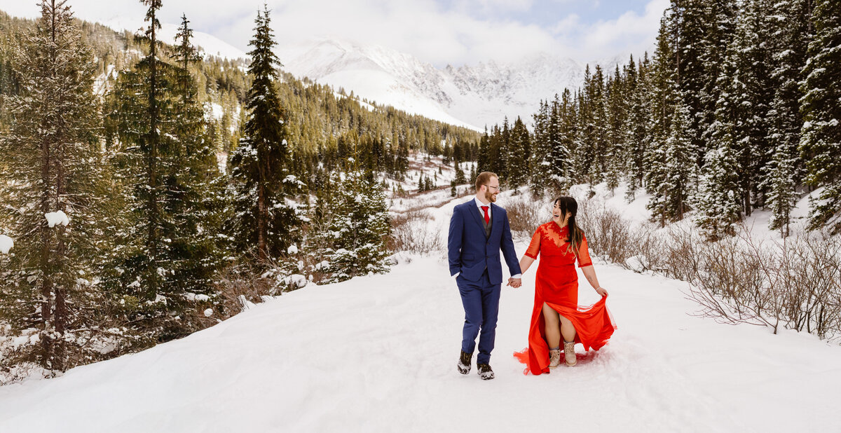 Couple runs through the snow hand in hand in the Colorado mountains while the bride wears a red dress during their Colorado elopement with Alpine Vows