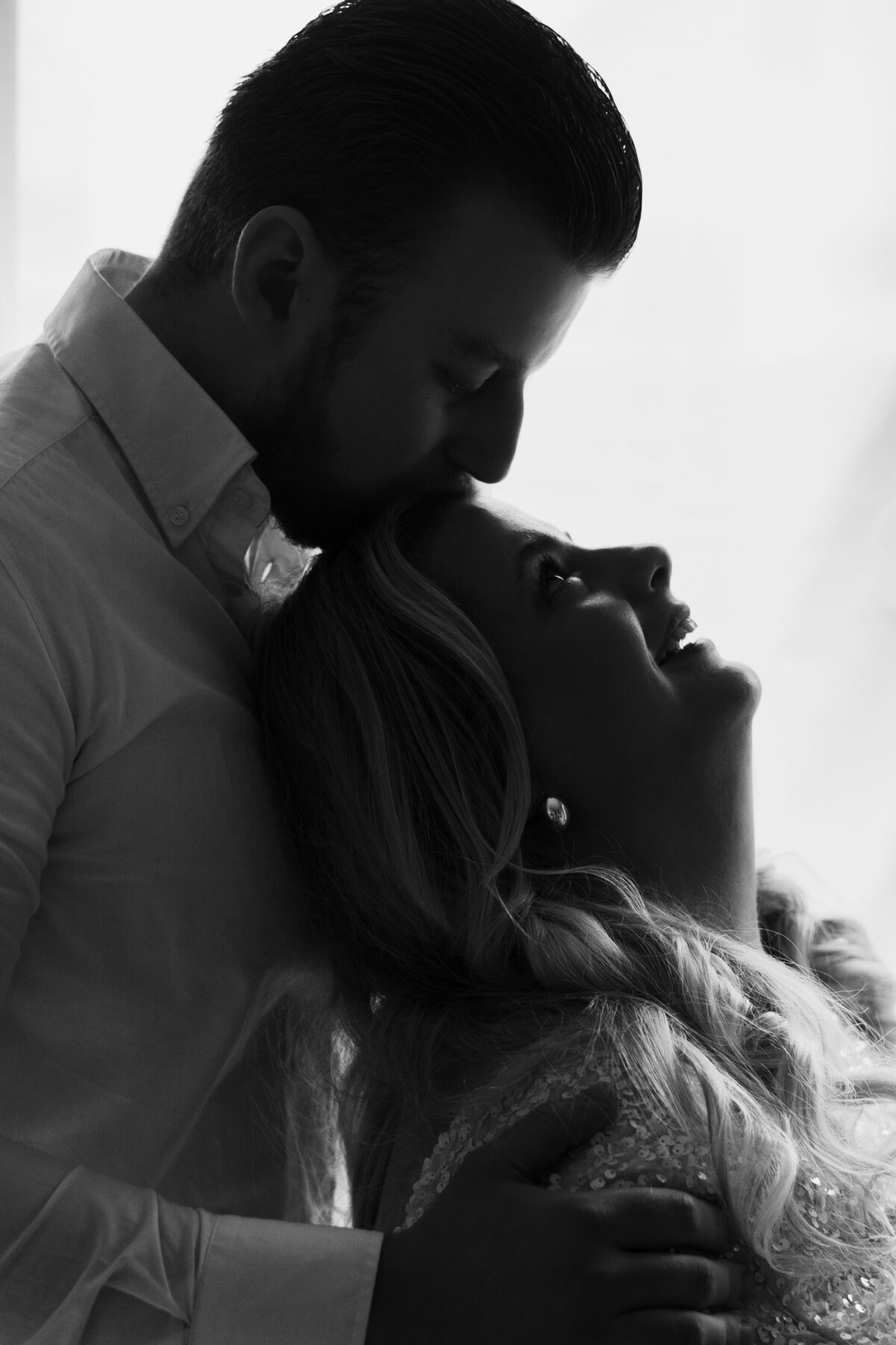 Een man en vrouw omarmen elkaar liefdevol voor een raam, met een warme sfeer en natuurlijk licht dat naar binnen valt.