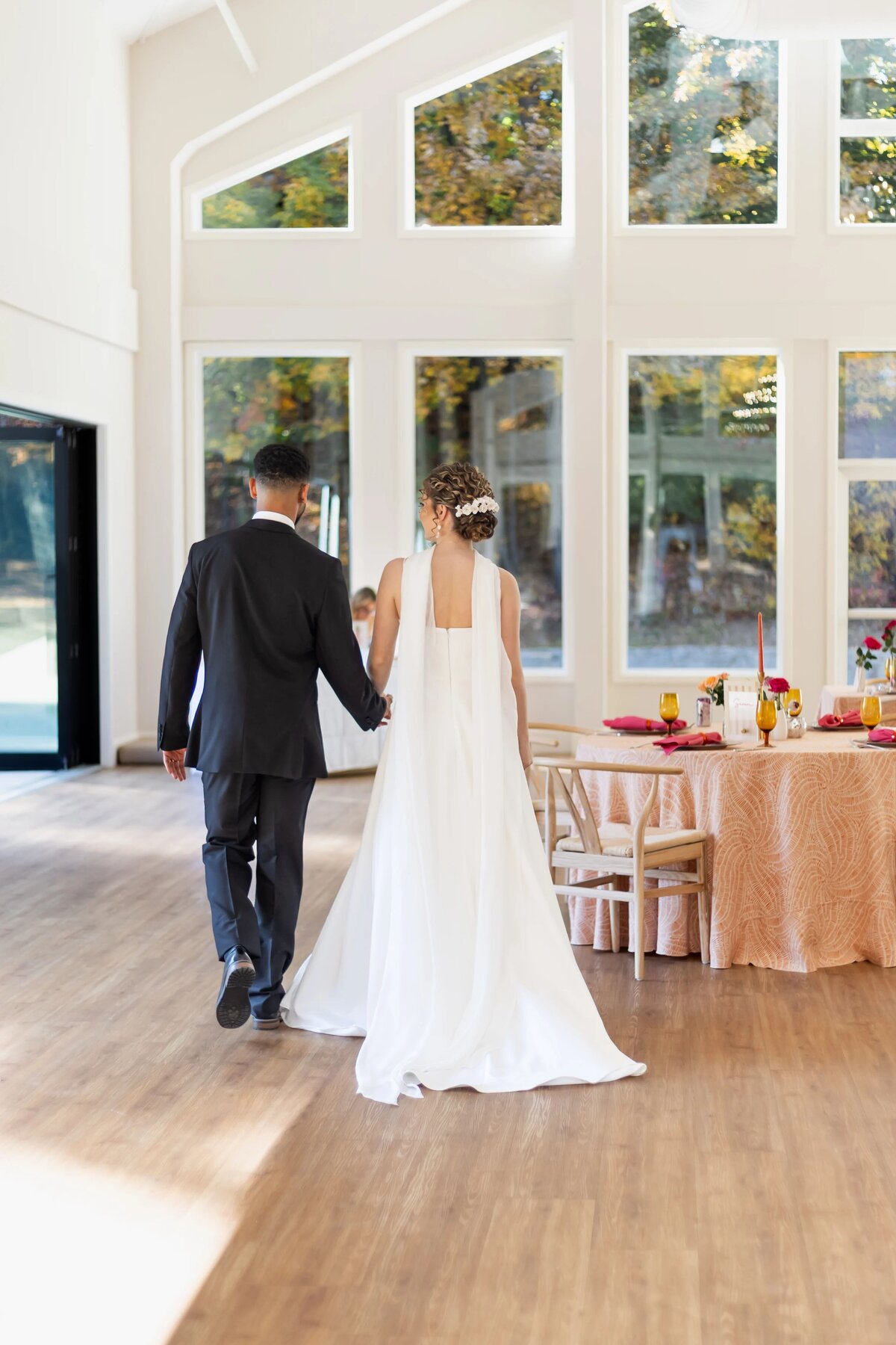 Wedding Photographers in RI | Kelsey Sheehan Photography Timeless Rhode Island Weddings | A bride in a white gown and a groom in a black suit walk hand in hand in a sunlit room with large windows, passing a table set for a celebration.