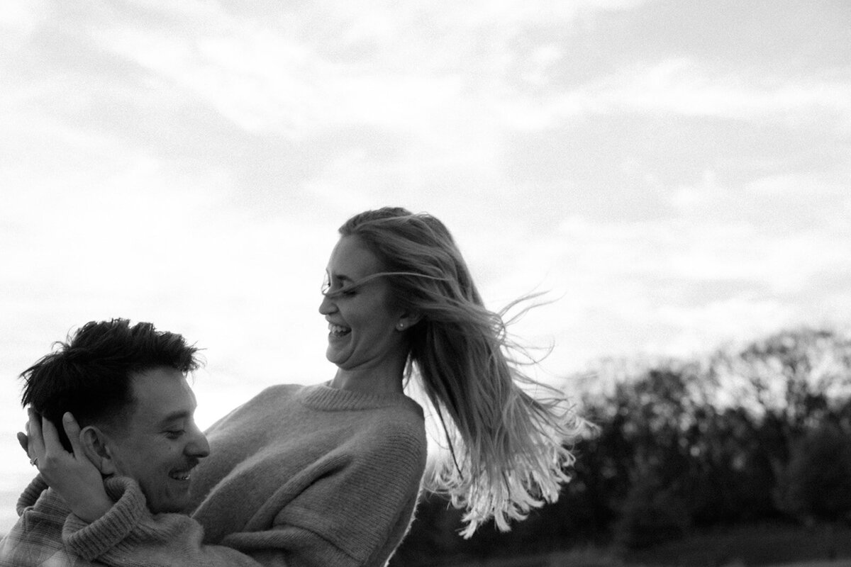 Couple smiles at each other while he spins her in the air and her hair waves in the wind. 