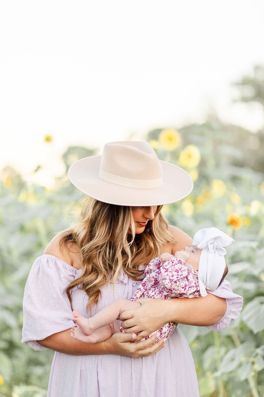Mom in purple off shoulder dress  wearing a light brown hat while holding baby who is wearing a purple flower outfit