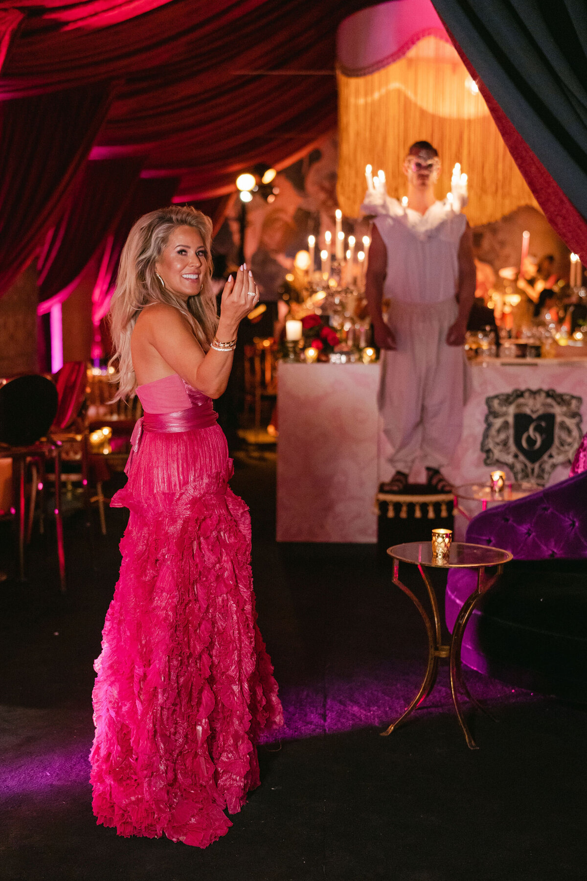 The stunning photo was captured in Virginia at a tented wedding by the talented photographer Elisabetta Marzetti, with Clayton Films as videographers. In the heart of this lavish event, a woman in a vibrant pink gown stands clapping, adding a pop of color against the backdrop of rich red and purple curtains. The ambiance is truly opulent, with the warm glow of candles, sparkling chandeliers, and intricate decor creating a magical atmosphere. As the scene unfolds, a figure in a white outfit can be seen standing on a platform in the background, adding to the overall elegance of the setting.