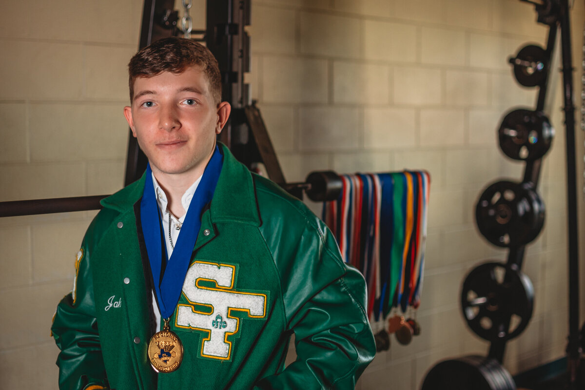 A Santa Fe High School senior boy stands near a weightlifting rack with his powerlifting medals in the background.