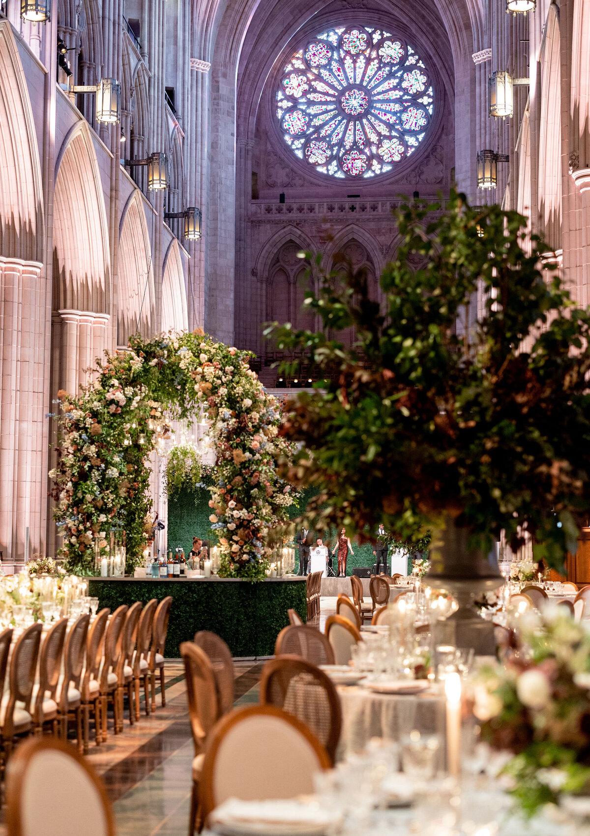 This is where the photo was taken at Washington D.C., Washington National Cathedral by photographer Twah Dougherty. The elegantly decorated cathedral setting features high vaulted ceilings and a large stained glass rose window, creating a breathtaking backdrop. The aisle is adorned with a lush floral arch, adding a touch of natural beauty to the space. Rows of tables dressed in white linens are surrounded by wooden chairs, elegantly set for a banquet or event, creating a welcoming and sophisticated atmosphere. Additionally, Current Films serves as the videographers capturing the special moments of the event.