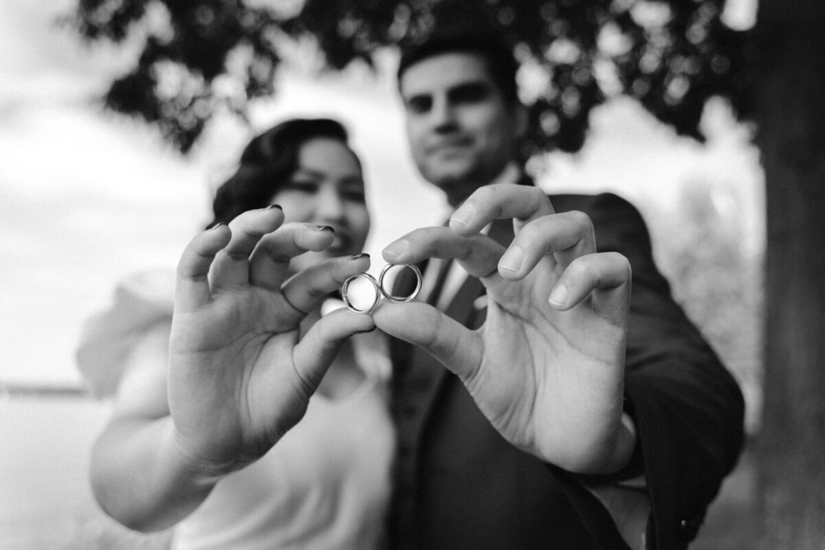 Black-and-white portrait of a bride and groom holding their wedding rings towards the camera, with their faces softly blurred in the background.
