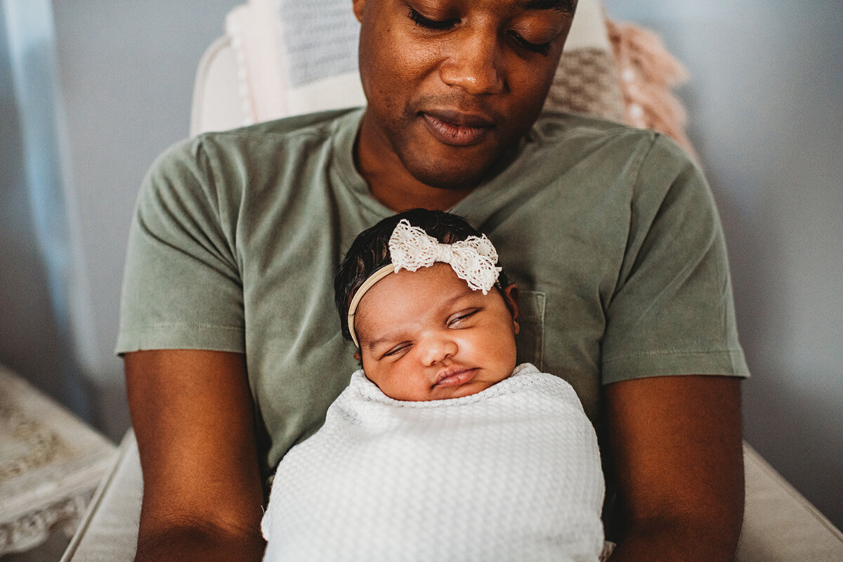 newborn photography with father sittiing in a nursery and holding his newborn daughter for family photos with a baby