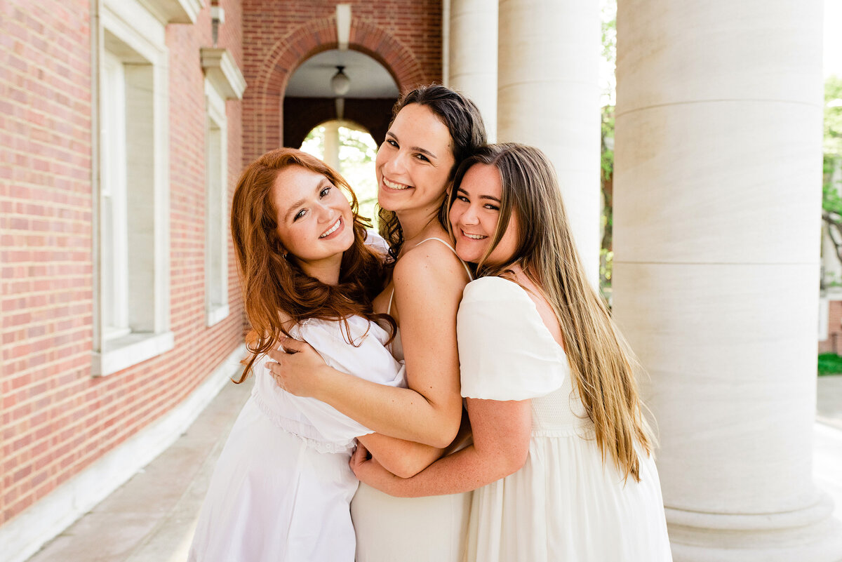 3 senior girls wearing white dresses and hugging one another while smiling at the camera