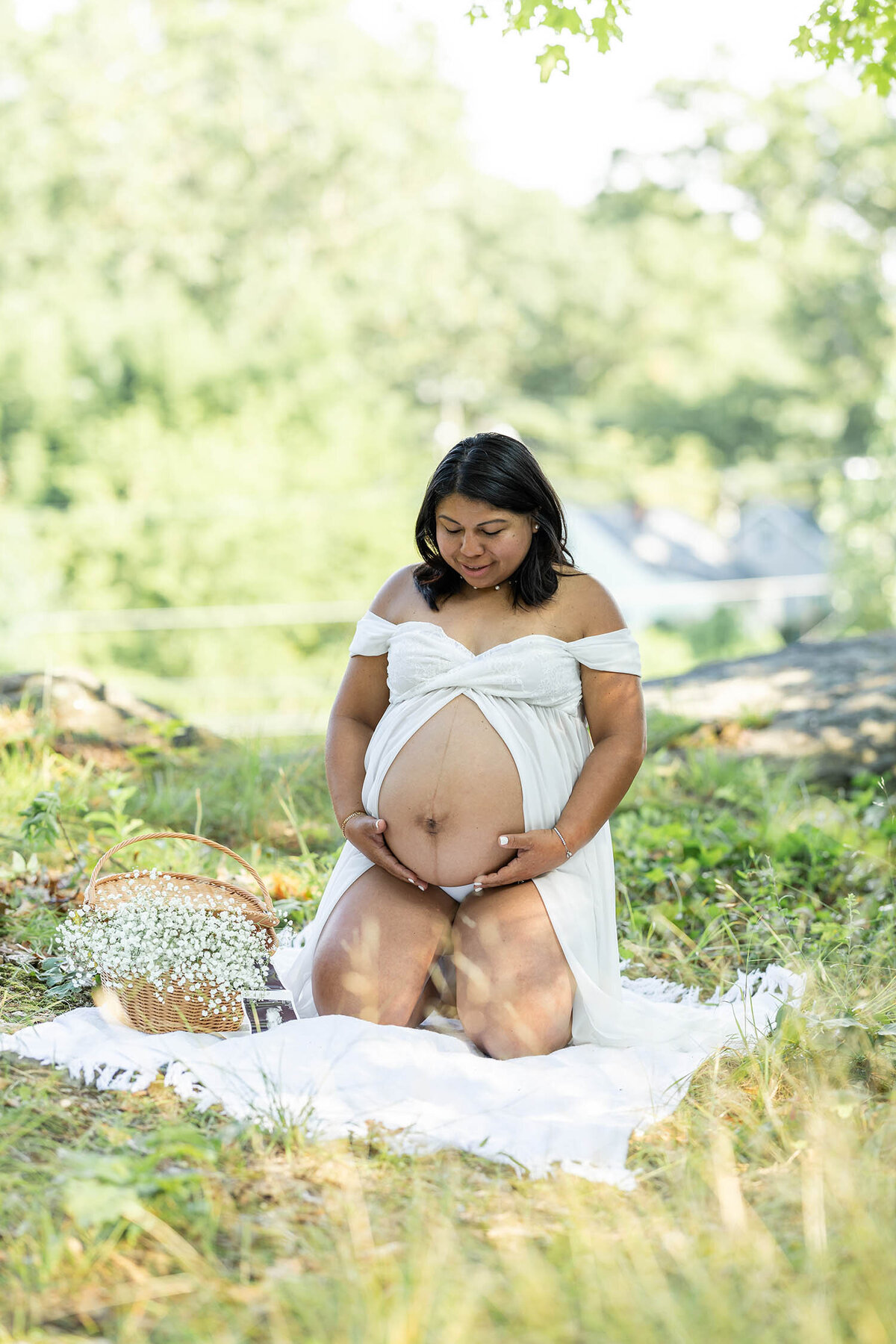 Pregnant woman wearing an open belly white dress kneels down as she looks down at her bump during her Maternity session in Connecticut