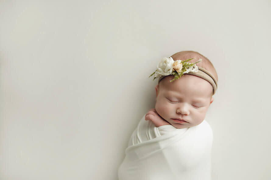 Baby with flower headband sleeps on a white backdrop for her photo session.