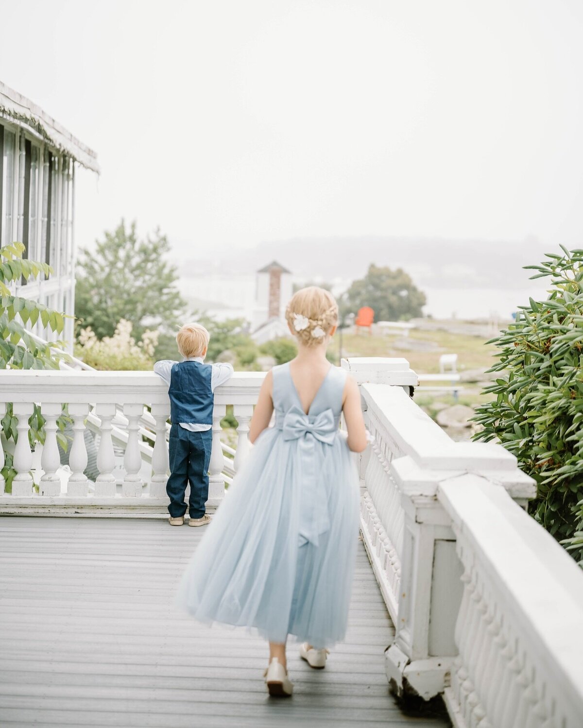 Wedding Photographer RI | A young girl in a light blue dress with a bow walks on a veranda, while a boy in a vest stands at the railing, overlooking a foggy landscape.
