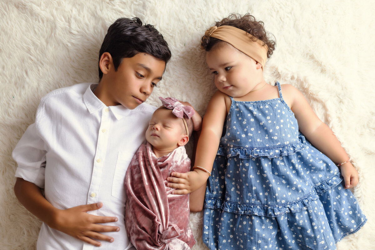 Newborn Photographer, a baby girl lays between her older brother and sister as they admire her
