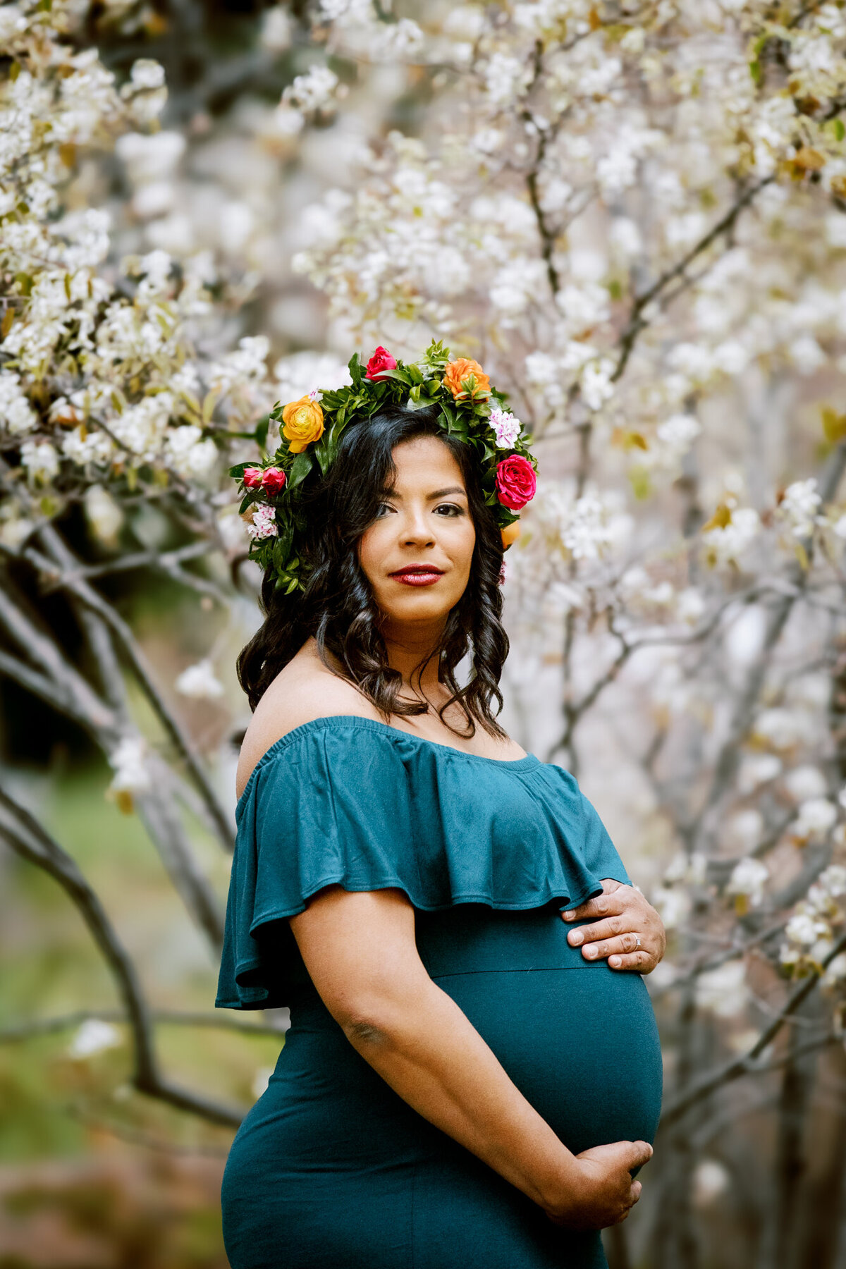 a pregnant woman in a turquoise dress and floral wreath holds her baby bump while standing in front of blooming trees at a park in Boulder, Colorado