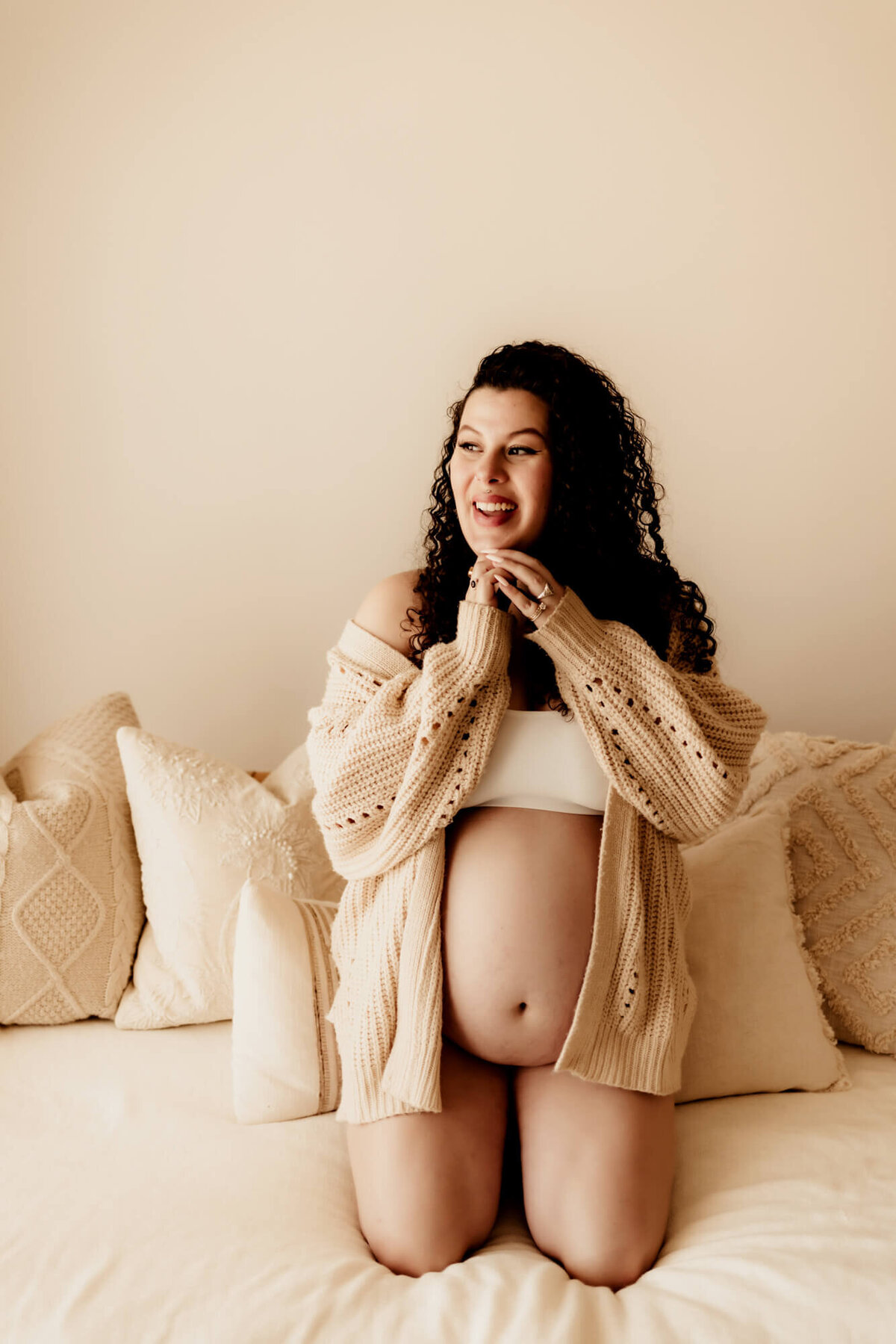 Pregnant woman wearing a beige sweater smiles with her hands under her chin. 