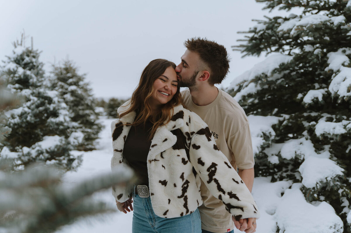 A woman leans back against her boyfriend as they hold hands. He's kissing her forehead as they stand in a snowy forest.