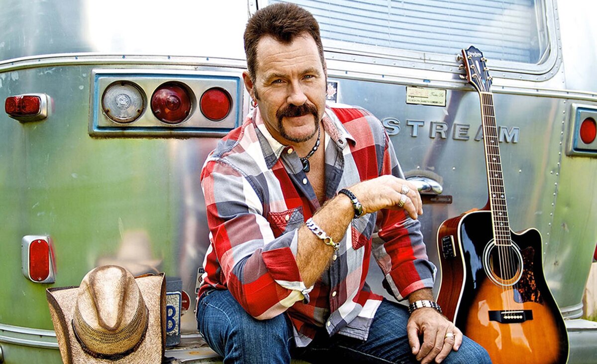 Country musician portrait Dean Young wearing plaid shirt sitting against old trailer with guitar resting beside