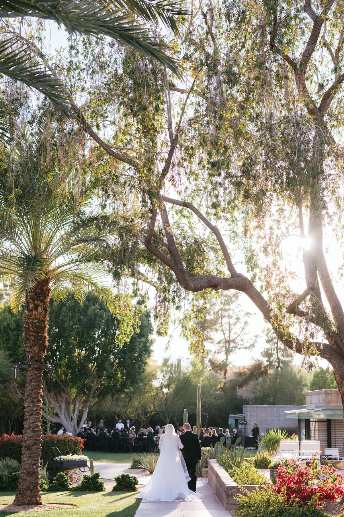 Bride walking down the aisle with her father during an outdoor ceremony at the Arizona Biltmore, surrounded by palm trees and desert gardens.