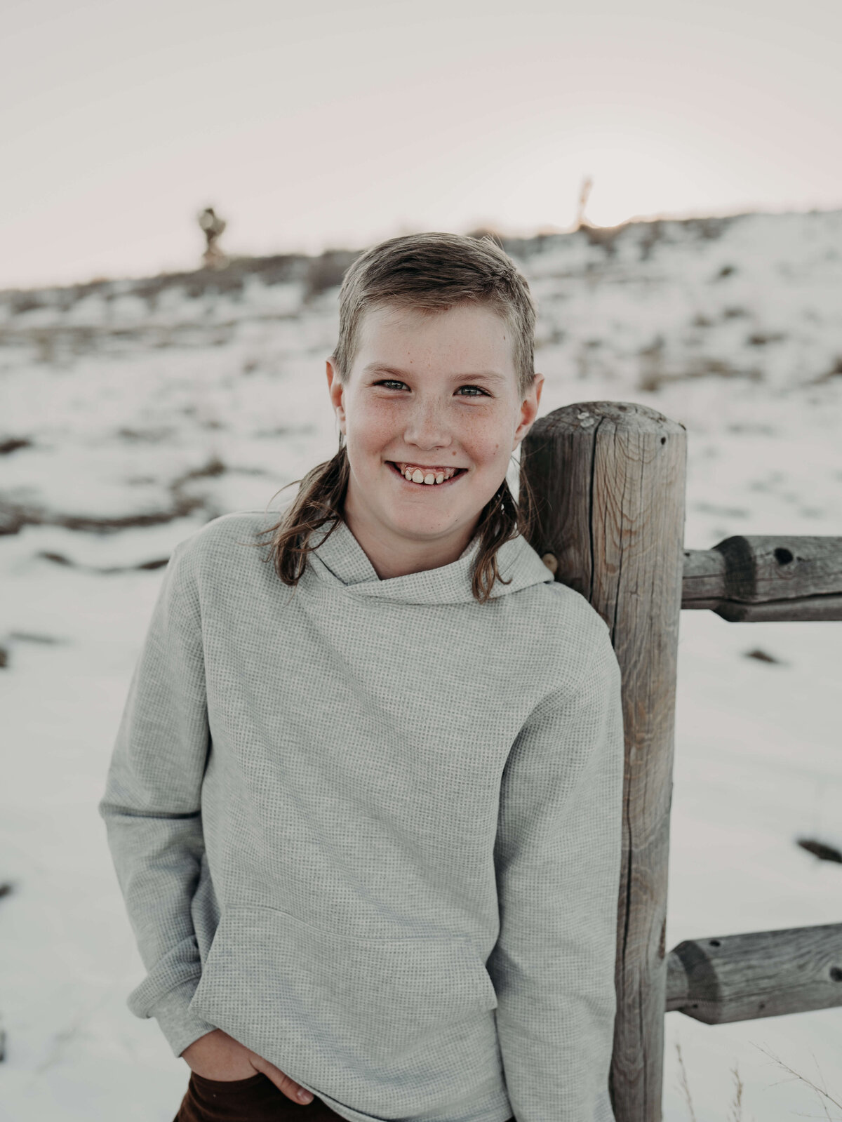 A close-up portrait of a 10-year-old boy, sporting a mullet and smiling at the camera with one hand in his pocket, as the snowy background complements his fair skin tones.