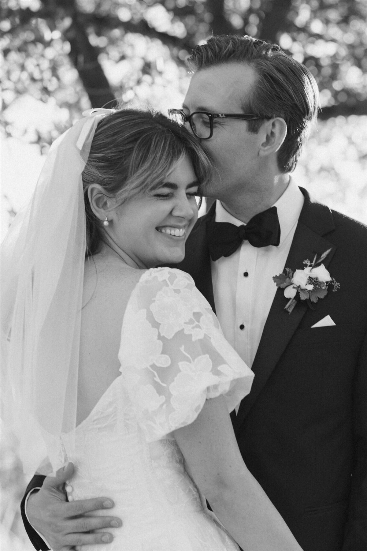 Bride is smiling at the camera while groom is kissing the brides head in black and white