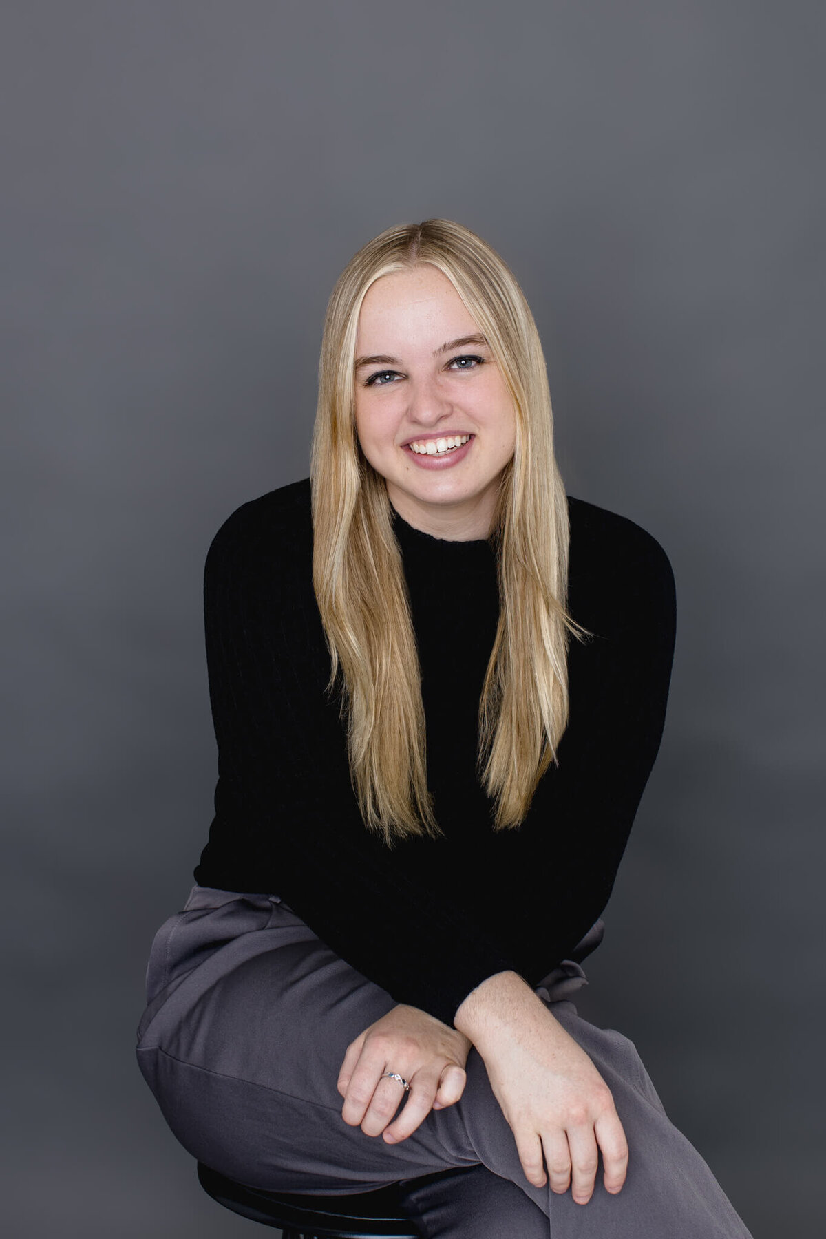 A young woman with long, blonde hair wearing a black turtleneck and grey dress pants sitting on a stool with her legs crossed at the knees and her arms on her knee, smiling at the camera in front of a grey background.