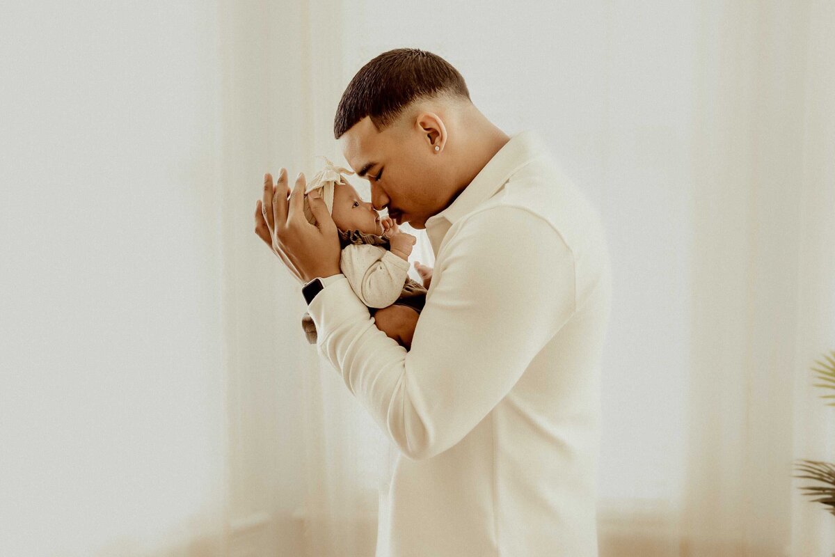 A man in a white shirt gently holds a newborn baby dressed in light clothing and a headband, looking down at the baby—a tender moment captured by Macon Newborn Photography.