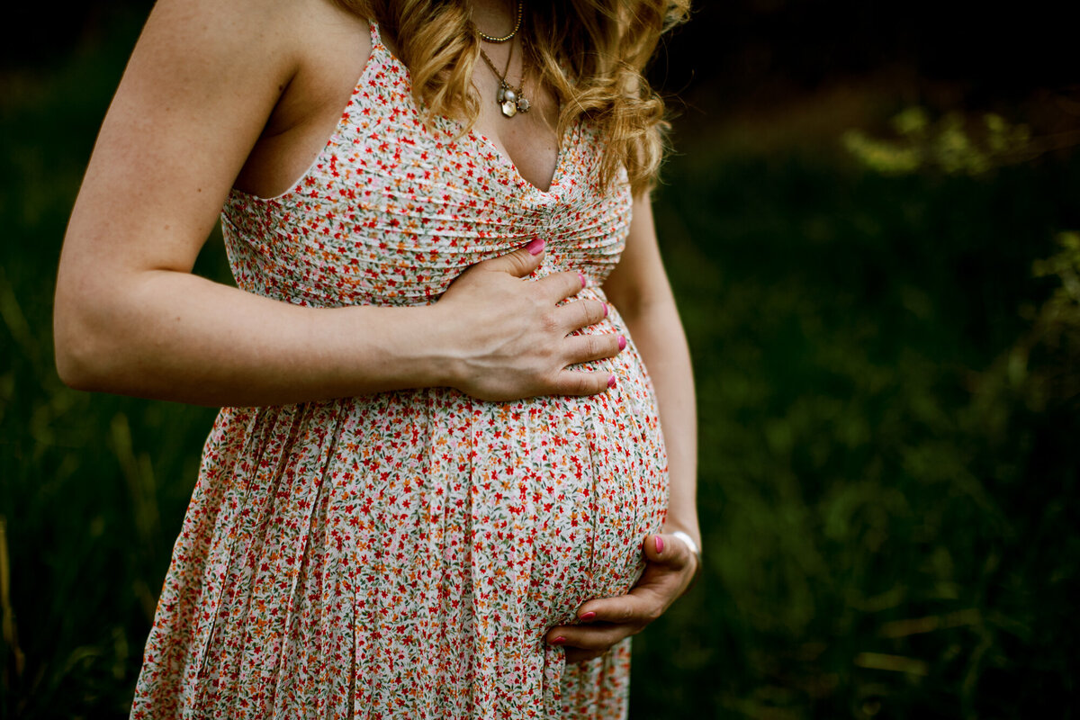 a woman in a floral dress holds her pregnant belly while standing in front of green grasses at a park in boulder, colorado