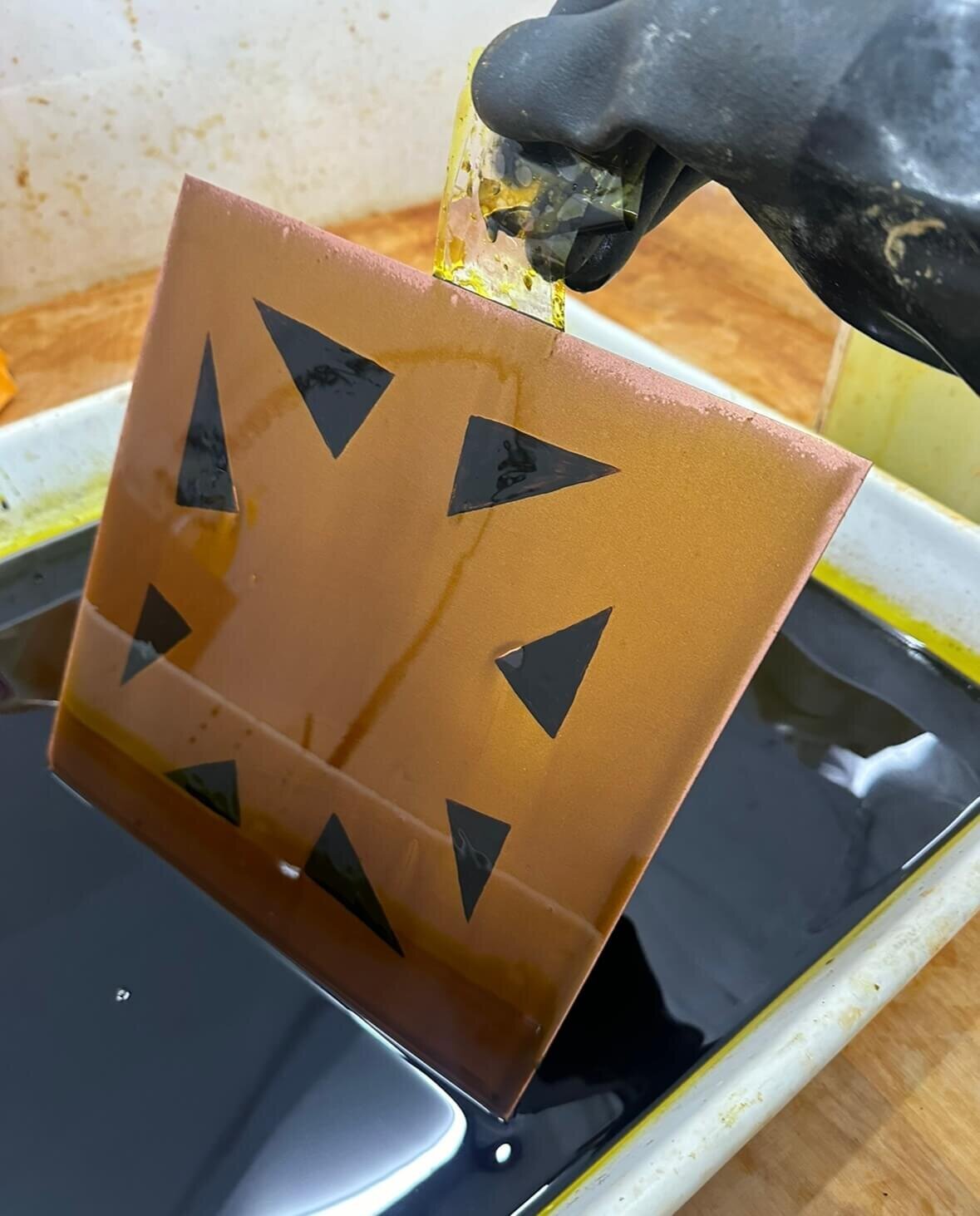 A triangular-patterned copper plate is lifted out of the acid bath with a gloved hand, mid-process in the etching sequence.