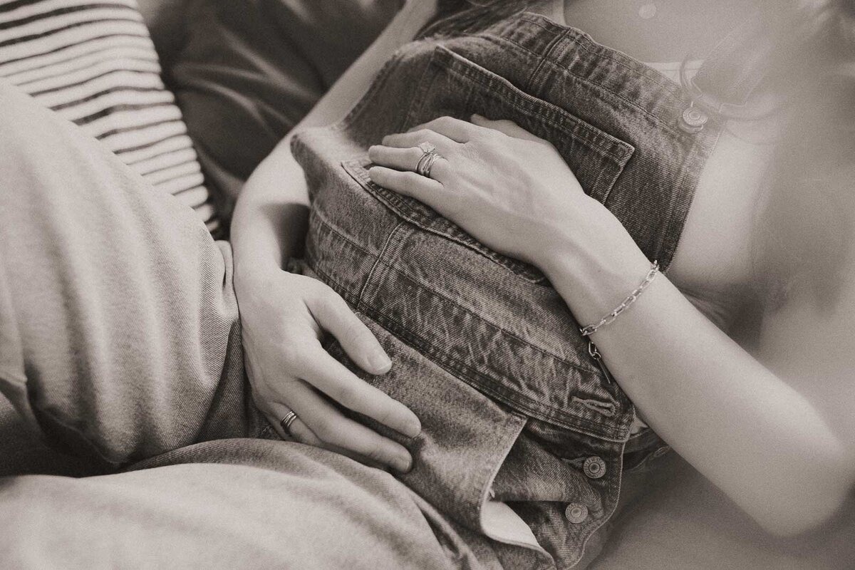 Pregnant person wearing overalls, lying down with hands resting on their belly, next to someone in a striped shirt.