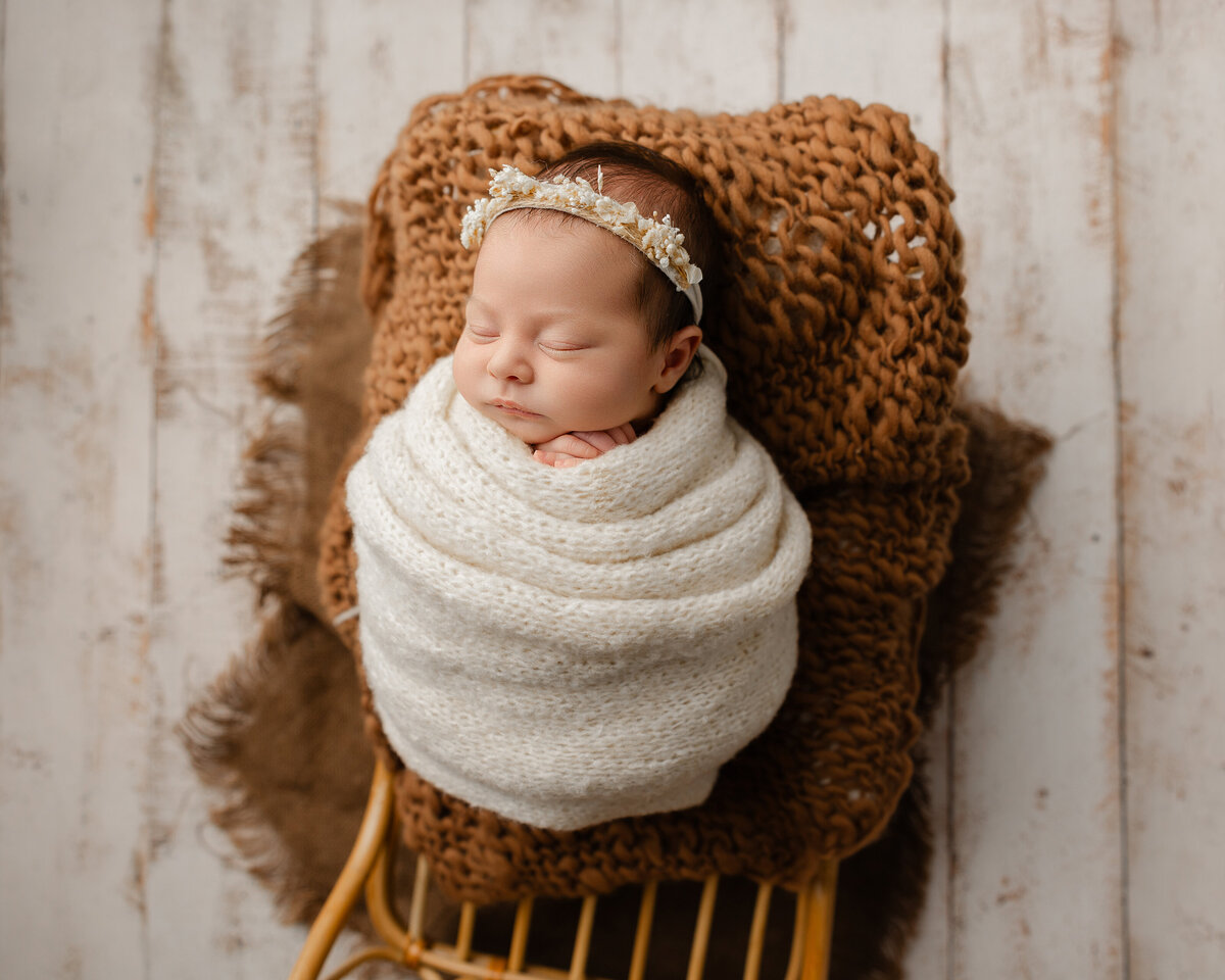 Newborn baby girl wrapped in soft ivory knits with a sweet floral crown, gently sleeping on a textured brown backdrop. Captured by Inland Empire newborn photographer Kaitlyn Dawn Photography during a cozy, serene session.