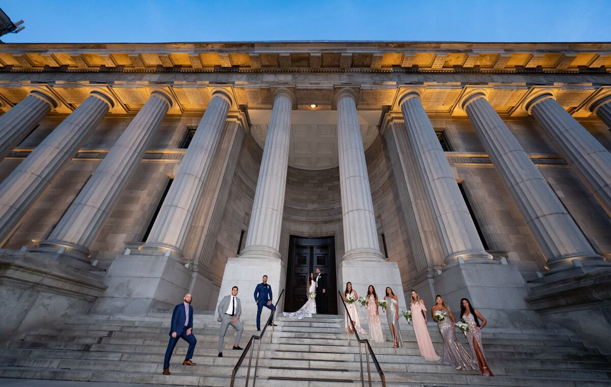 The bridal party stands on the steps of a majestic building with large Roman columns. They are dressed in formal attire, with the bride and groom prominently positioned among them. The grandeur of the building and the elegance of the group create a dramatic and sophisticated backdrop for this wedding portrait.