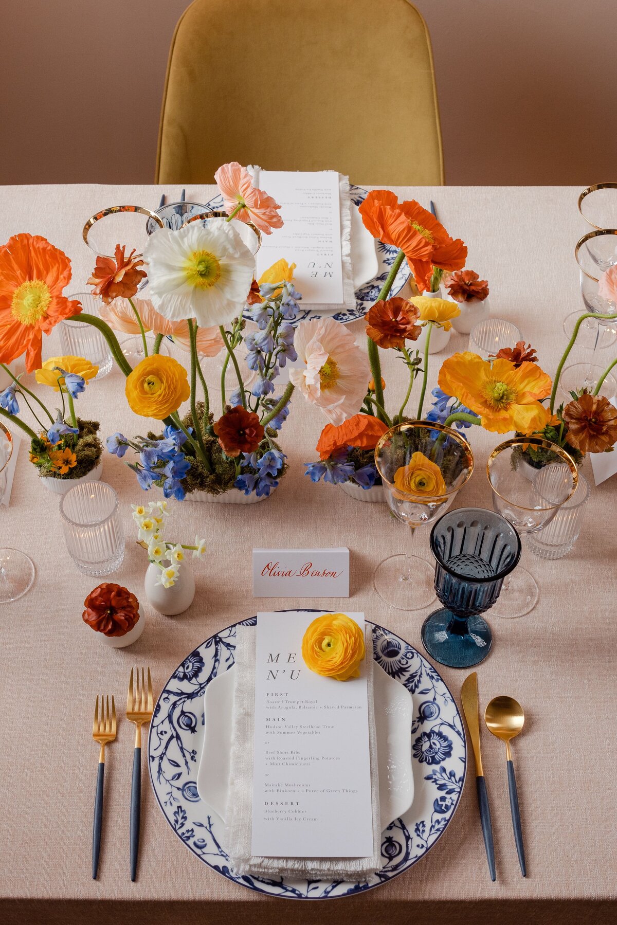 Wedding table setting with vibrant orange and yellow floral arrangements, blue glassware, and gold accents