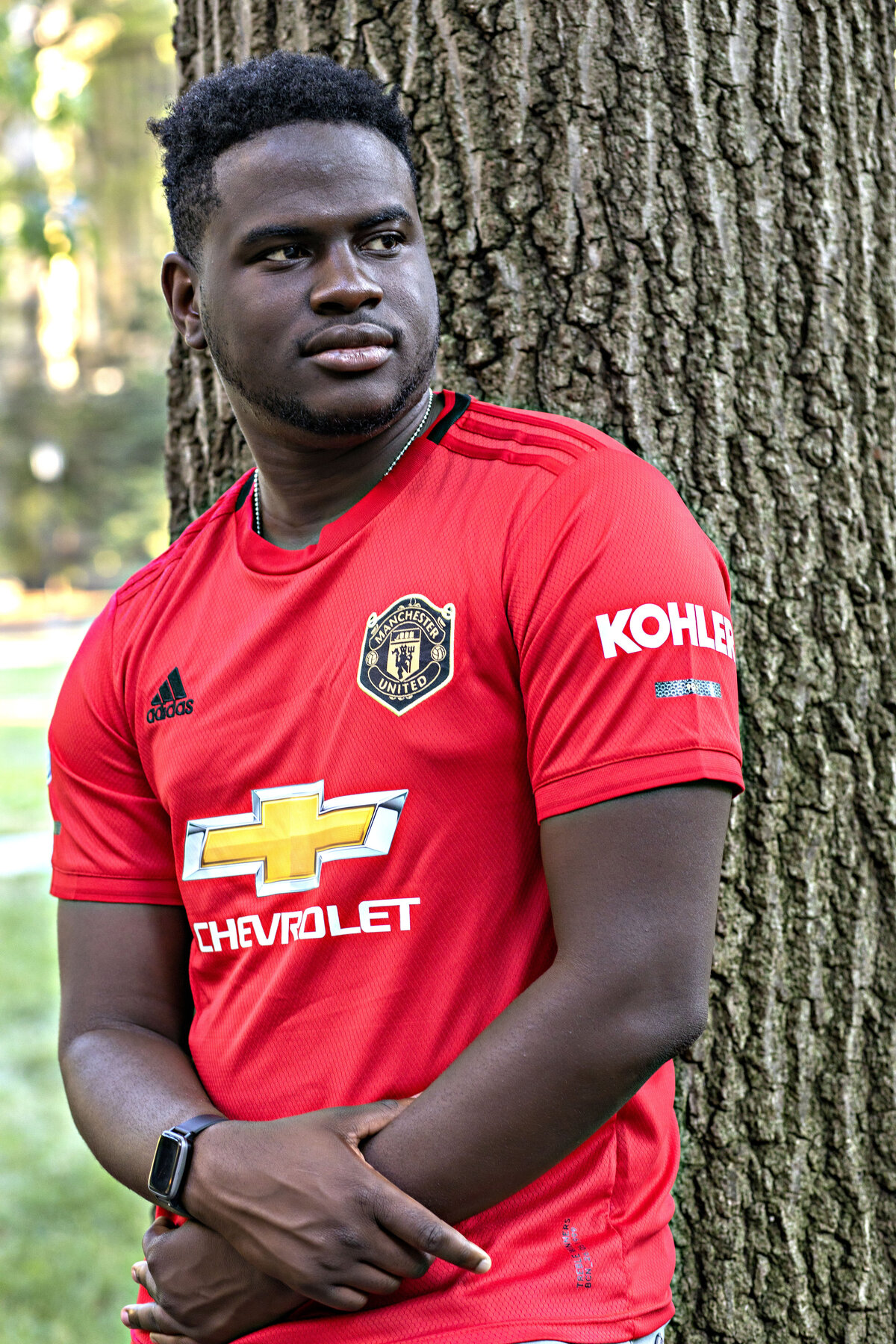 African American male wearing red sports shirt and grey jeans leaning against a tree