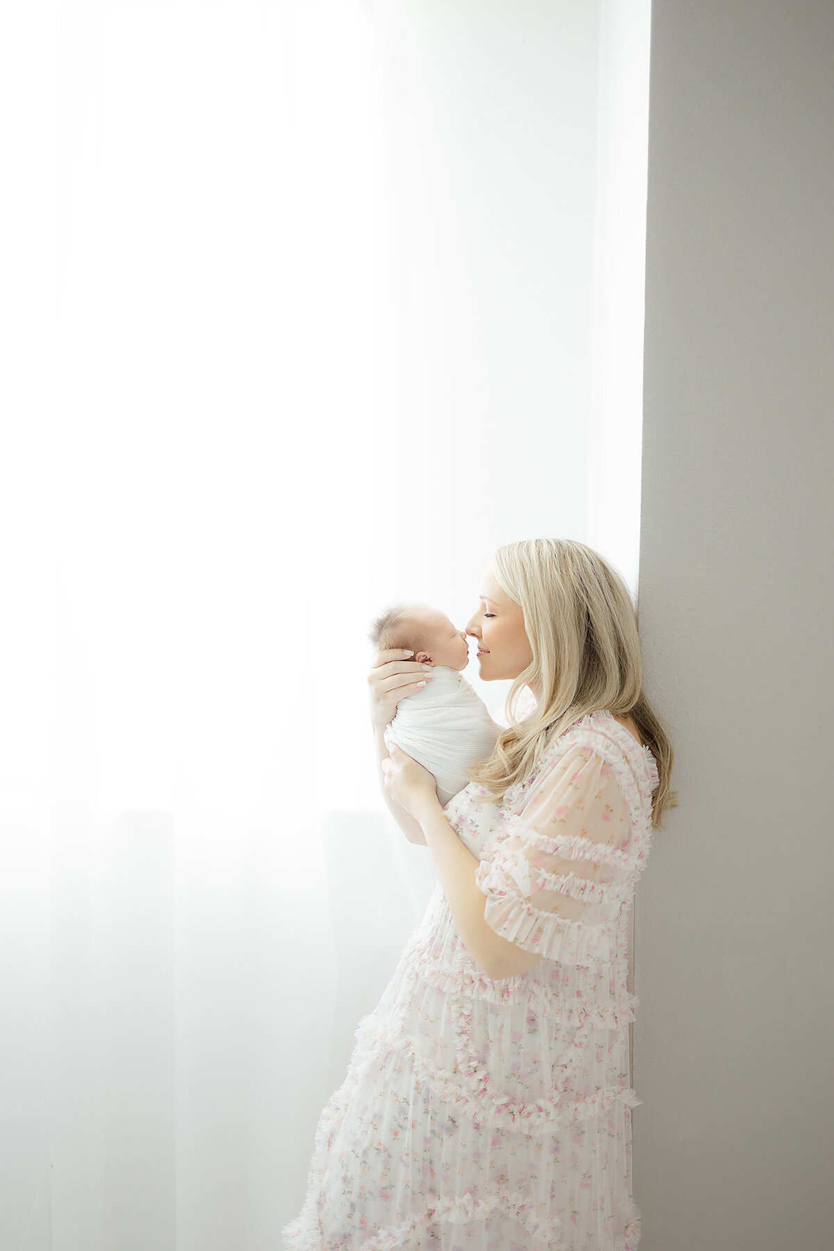 In studio newborn photo of a mother leaning against the wall while she holds her newborn baby girl close as they touch noses.